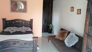 A bedroom with a bed, a sofa, a potted plant, and a wall with framed pictures.