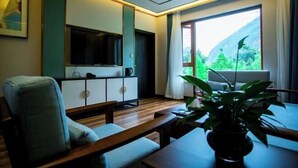 Een moderne woonkamer met een bank, een salontafel, een televisie en een groot raam met uitzicht op groen.