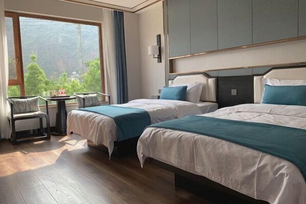 Een hotelkamer met twee bedden, een kleine tafel, een stoel en een groot raam met uitzicht op groen.