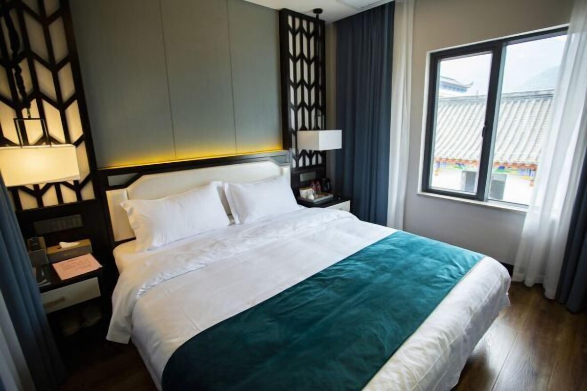 Habitación de hotel con cama, mesita de noche, lámpara y ventana con vistas.