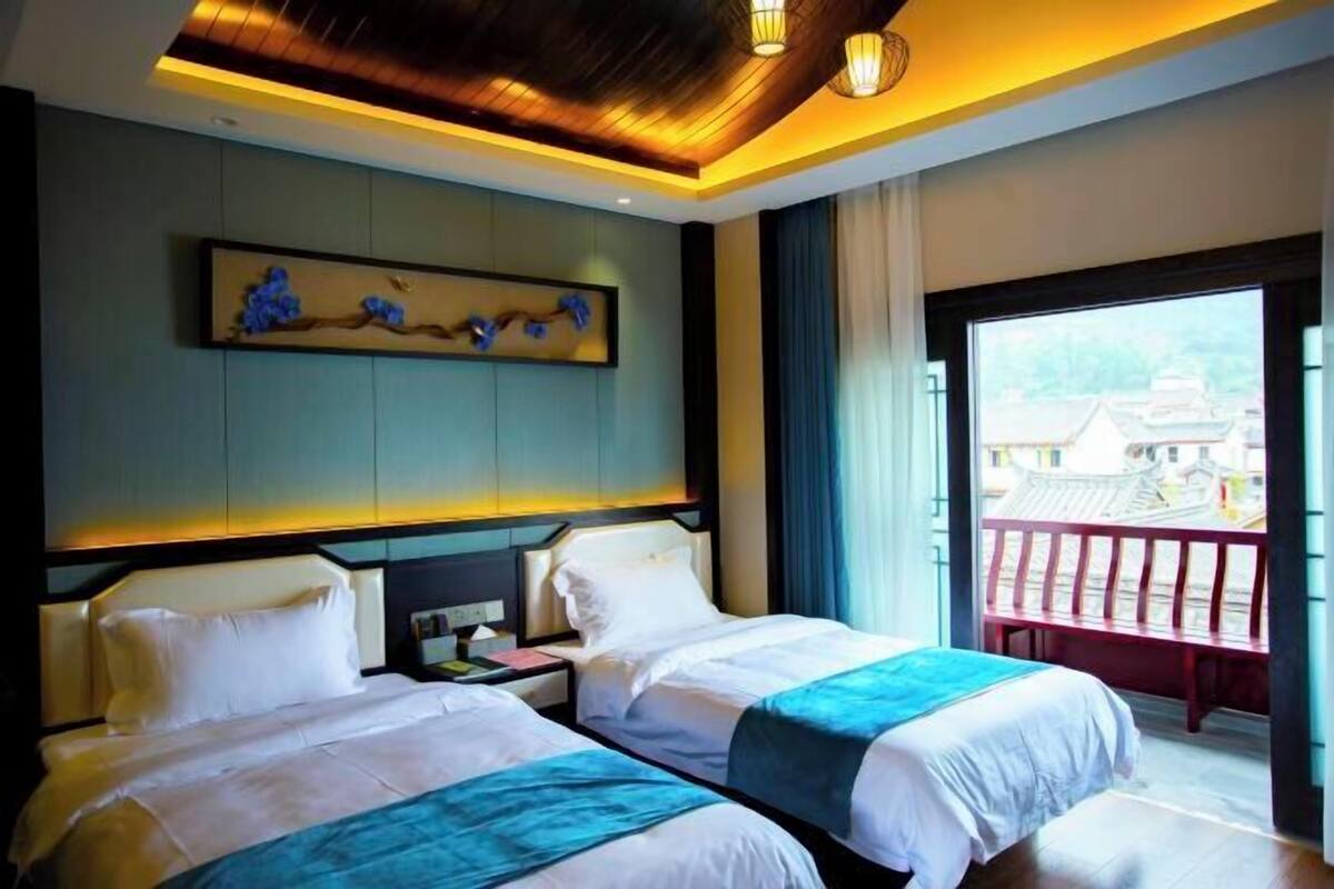 A hotel room with two beds, a large window with a view of traditional architecture, and a ceiling with recessed lighting.