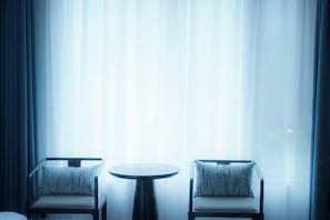 Una habitación con dos sillones y una pequeña mesa redonda, ubicada frente a un gran ventanal con cortinas ligeras.