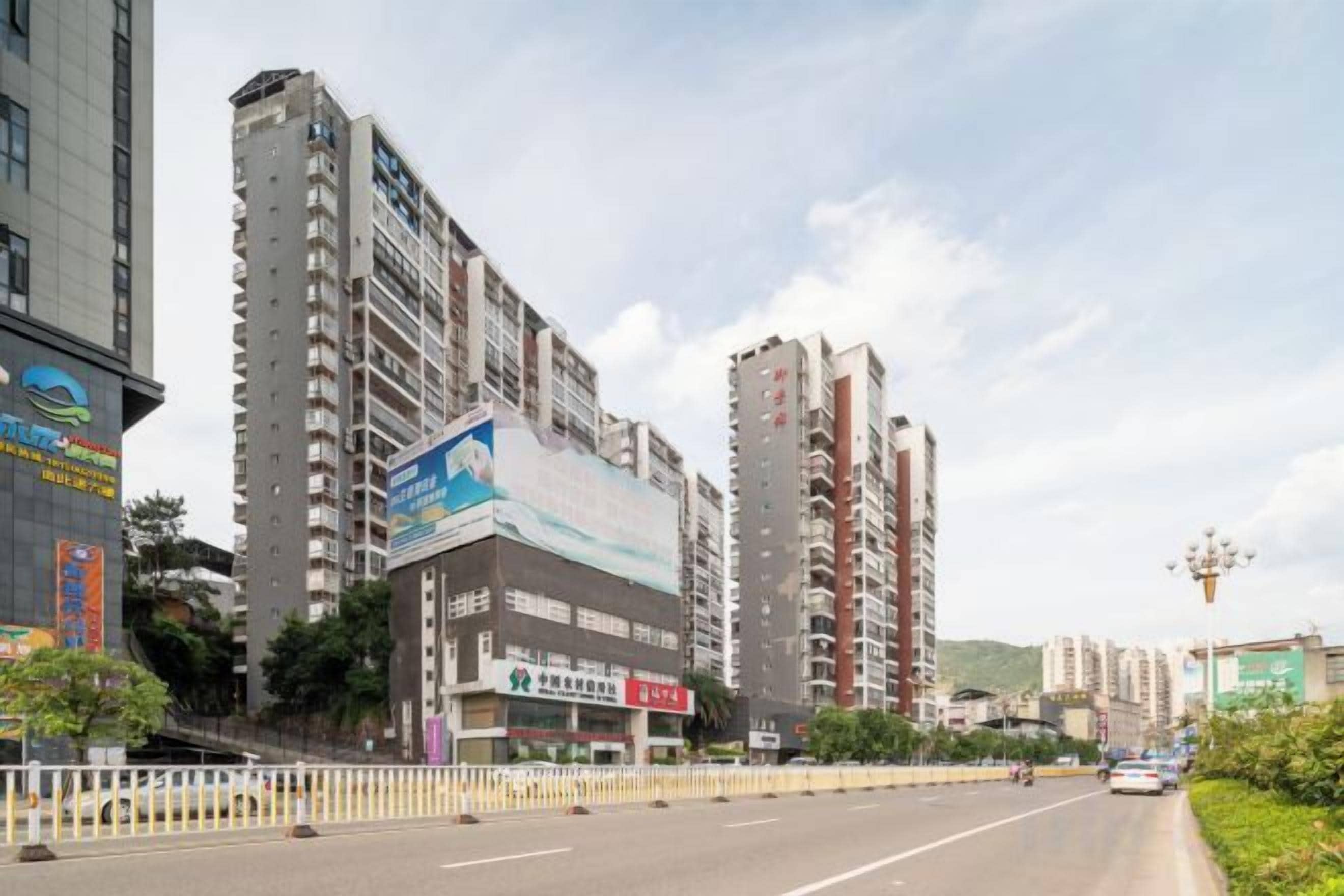 A multi-story building with a large billboard, a street with a white fence, and a clear sky.