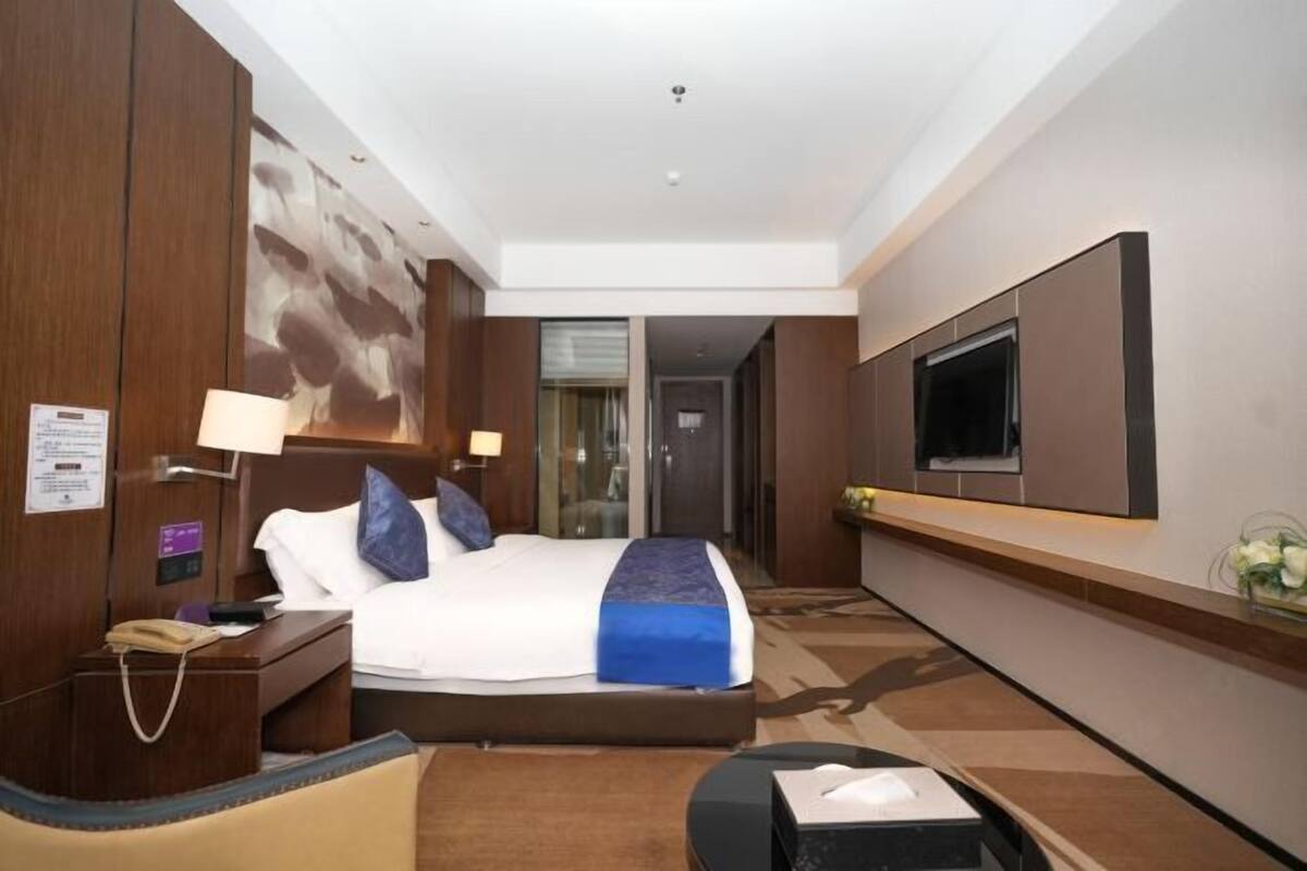 A hotel room with a large bed, a TV mounted on the wall, a desk with a lamp, and a small table with a tissue box.