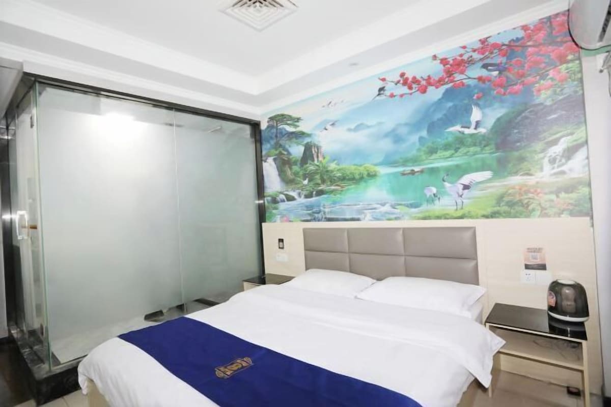 A hotel room with a bed, a glass shower enclosure, a mural of a landscape, and a nightstand with a kettle.