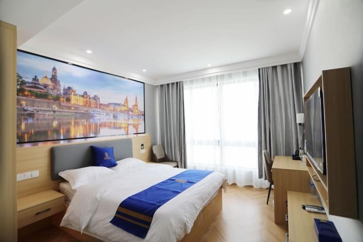 A hotel room with a large bed, a desk, a TV, and a painting of a cityscape.