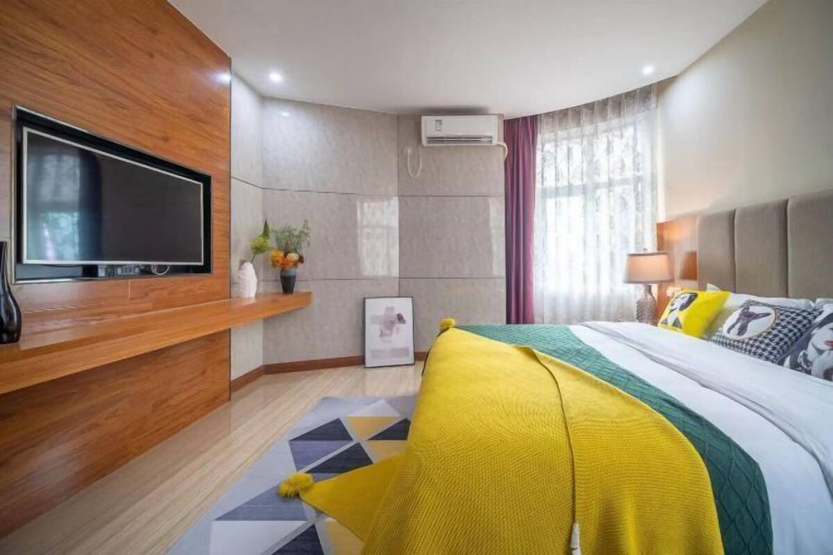 Een moderne hotelkamer met een groot flatscreen-televisie aan een houten wand, een bed met een gele en groene dekbedovertrek, een nachtkastje met een lamp en een airconditioner aan de wand.