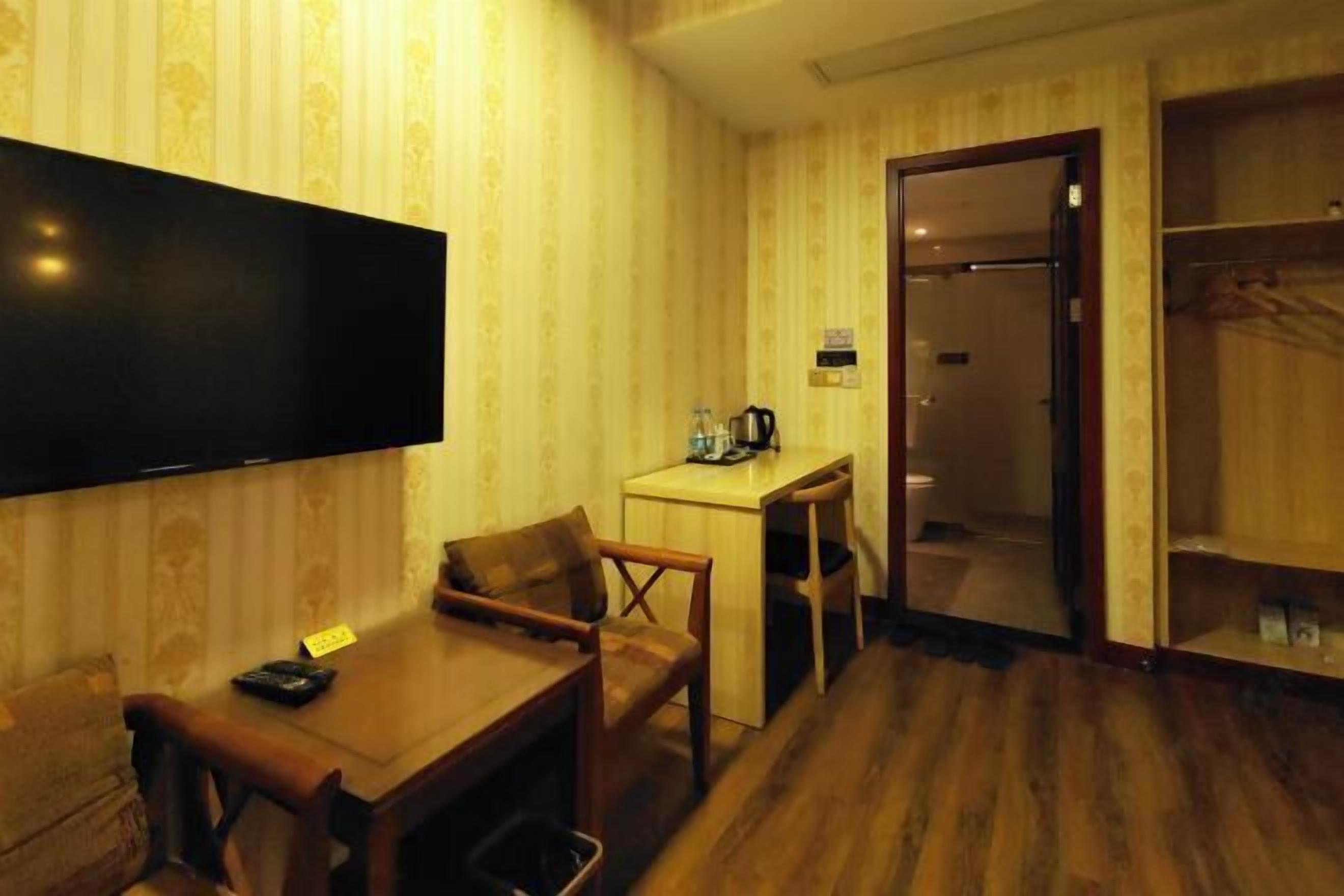 A hotel room with a wooden floor, a TV mounted on the wall, a small desk with a chair, and a wardrobe.