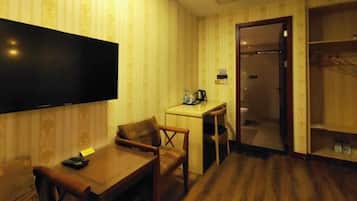 A hotel room with a wooden floor, a TV mounted on the wall, a small desk with a chair, and a wardrobe.