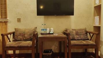 A room with two wooden chairs with patterned cushions, a small table with a kettle and bottles, and a flat-screen TV mounted on the wall.