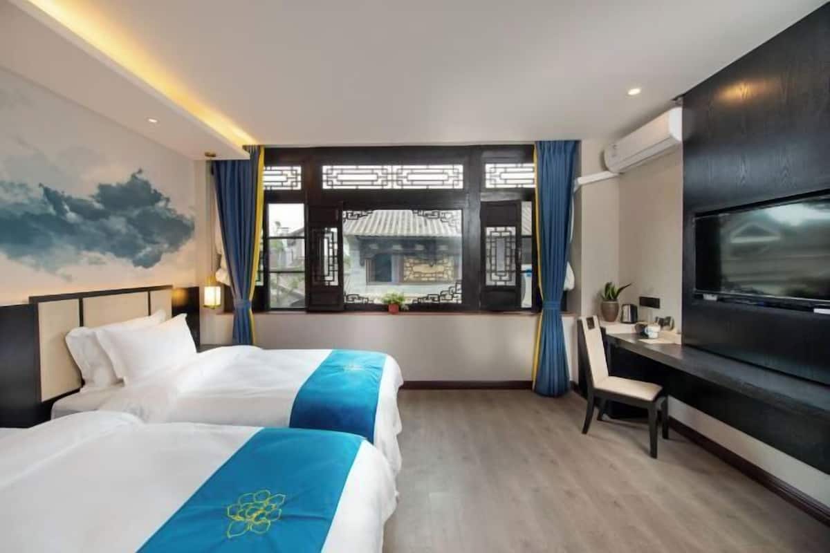 Habitación de hotel con dos camas, un escritorio, un televisor y una ventana que ofrece vistas a la arquitectura tradicional.