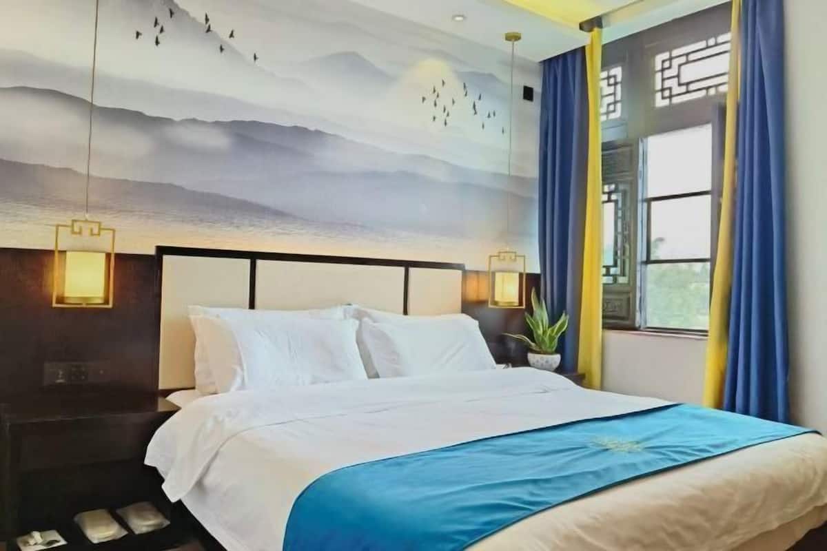 Habitación de hotel con cama, mesita de noche, un mural en la pared, ventana con cortinas y lámpara colgante.