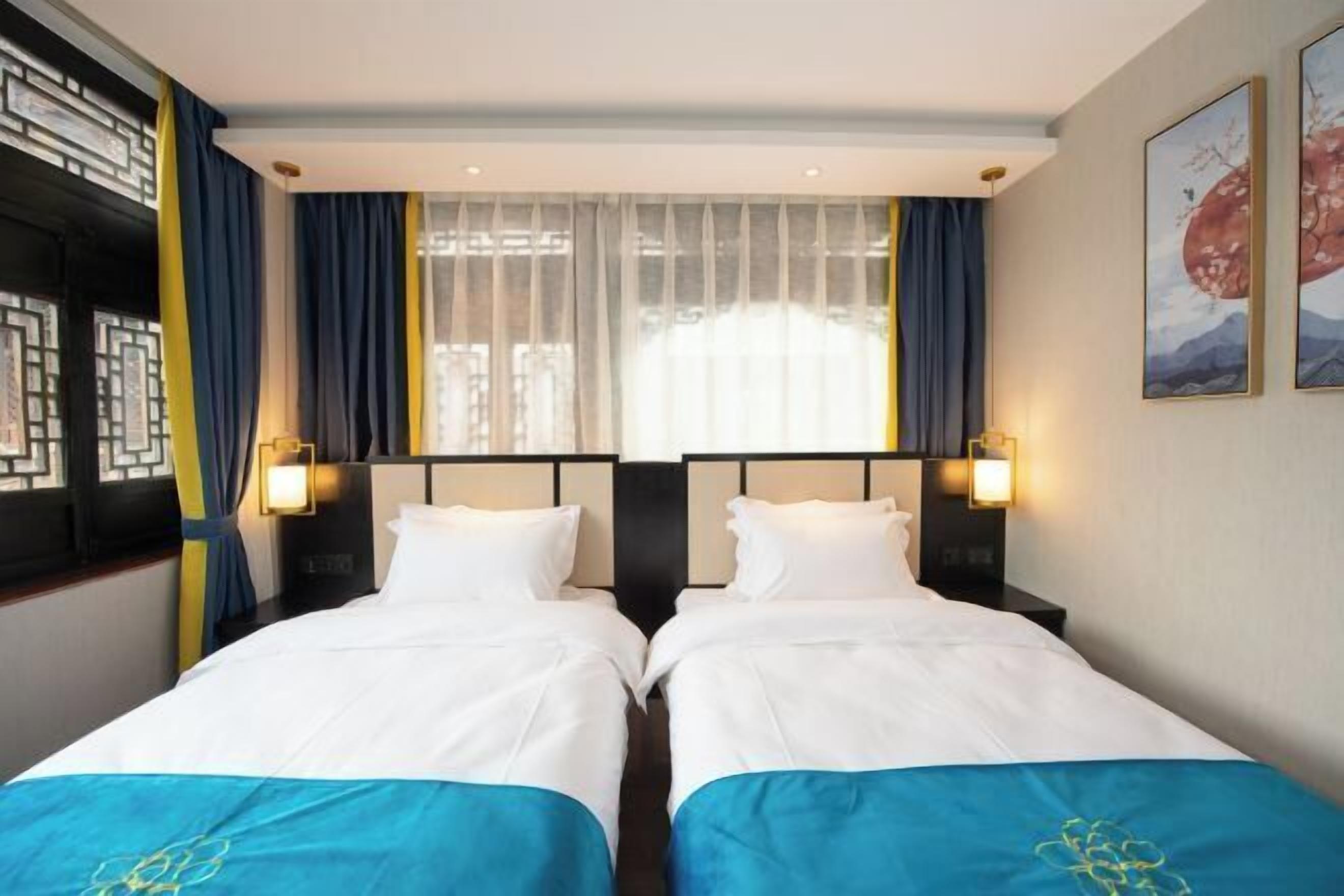 A hotel room with two beds, a large window with sheer curtains, and wall-mounted lamps.