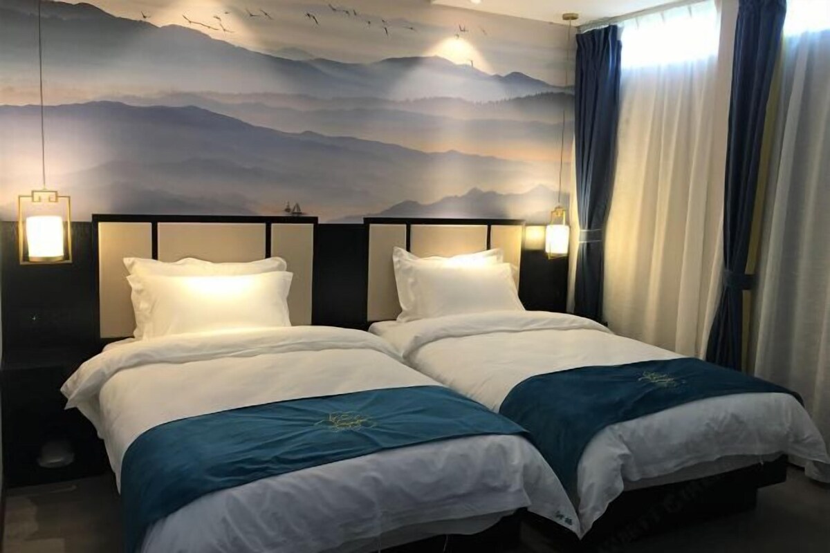 A hotel room with two beds, each with white bedding and blue accents. The room features a large wall mural depicting mountains and birds, and there are two bedside lamps.