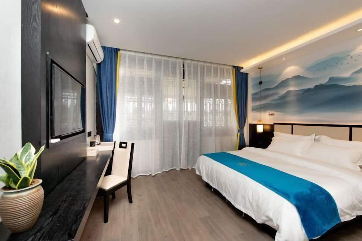 Una habitación de hotel con cama, escritorio, silla, televisión y un gran ventanal con cortinas translúcidas.