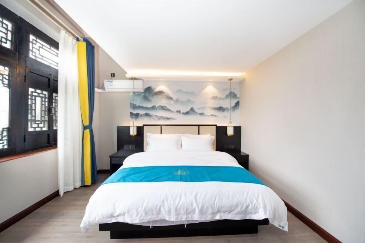 A hotel room with a bed, a wall-mounted air conditioning unit, and a painting of mountains.