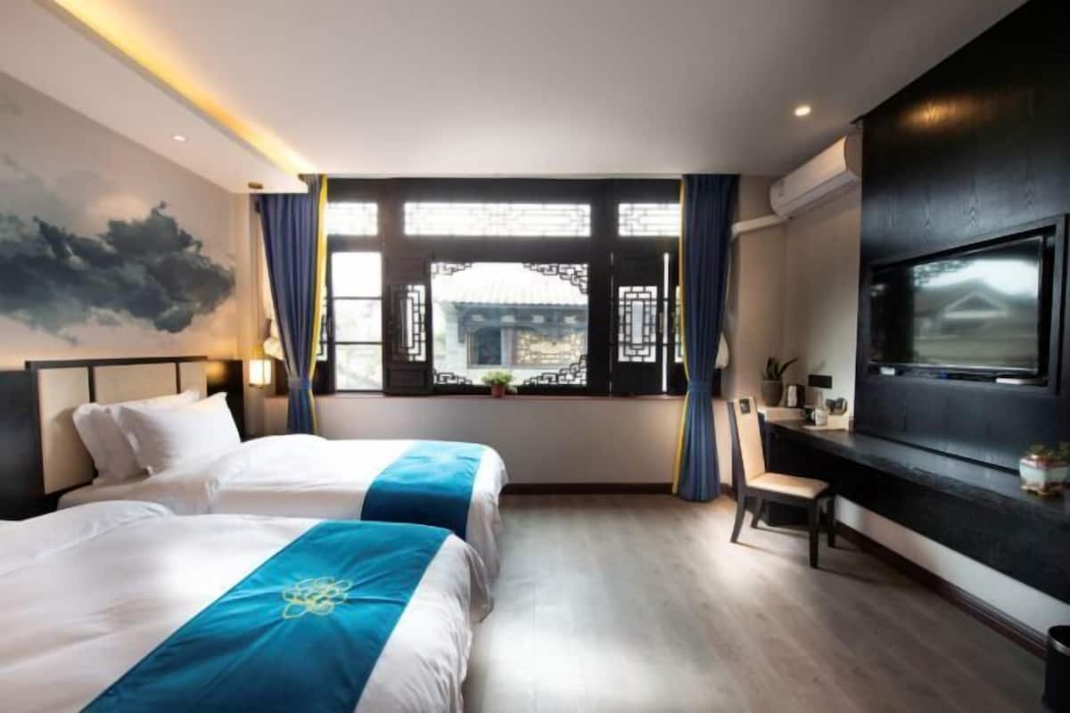Una habitación de hotel moderna con dos camas, un ventanal con diseño de celosía china tradicional, un televisor de pantalla plana montado en la pared y un escritorio con una silla.