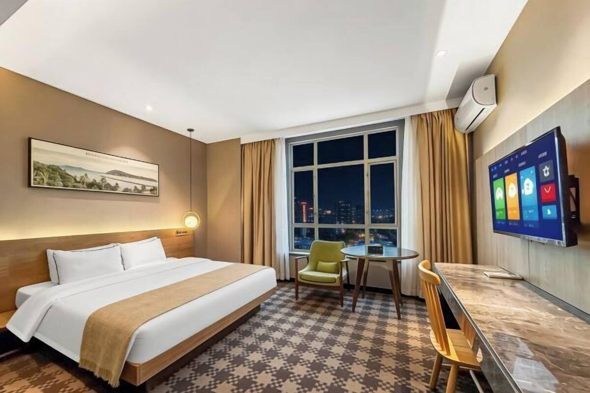 A hotel room with a bed, a desk, a chair, a TV, a window with a city view, and an air conditioning unit.