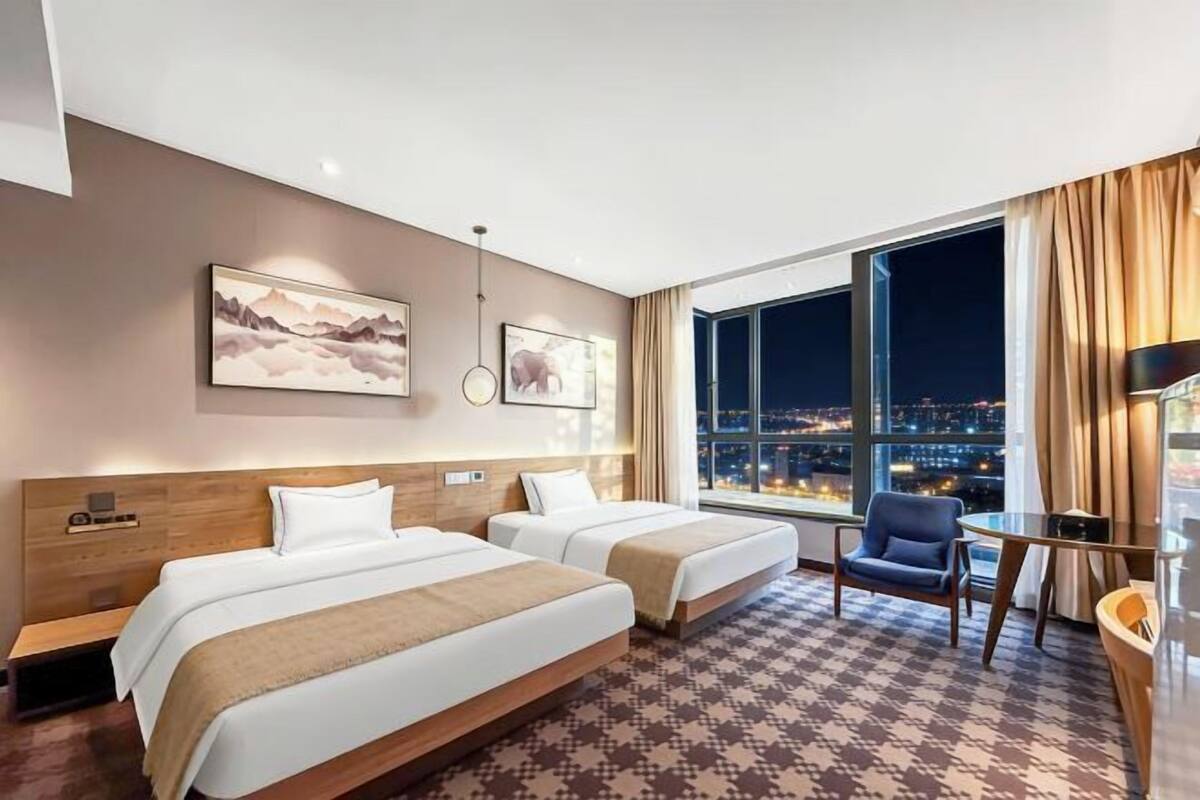 A hotel room with two beds, a desk, and a chair. There is a large window with a city view at night.
