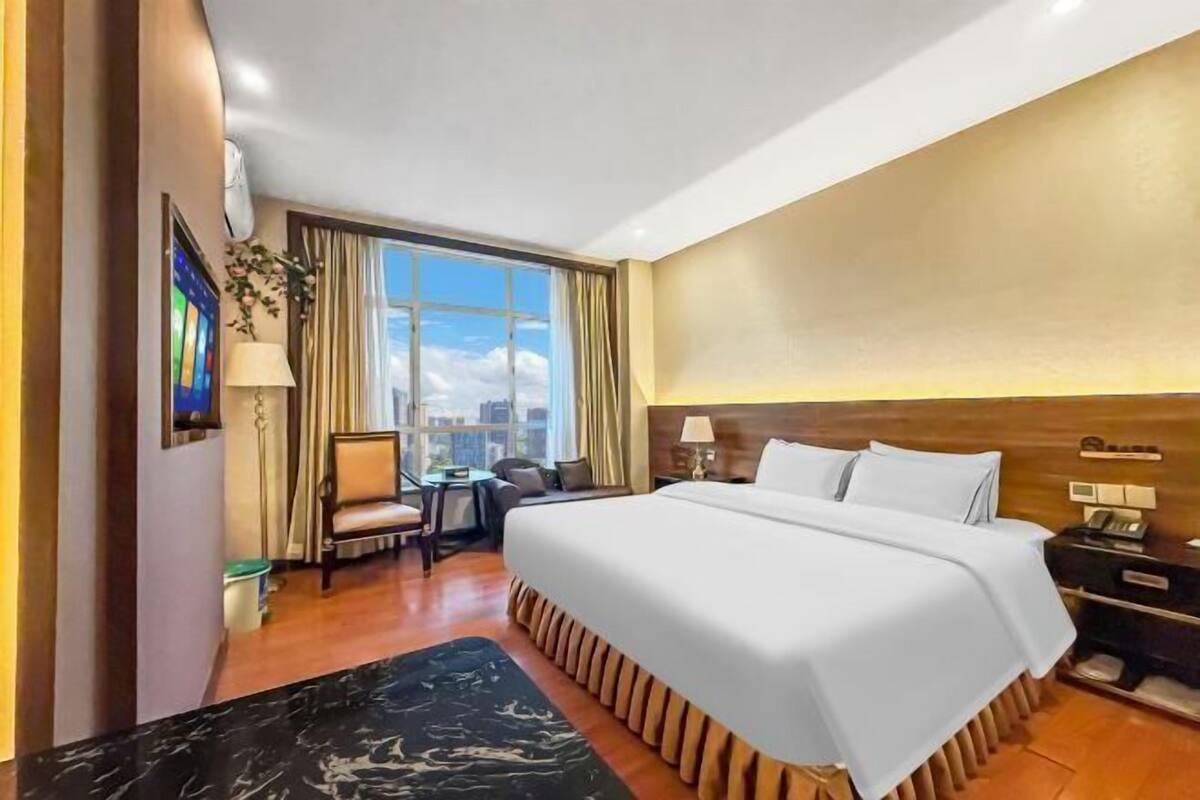 A hotel room with a large bed, a desk, and a chair. There is a window with a view of the city.