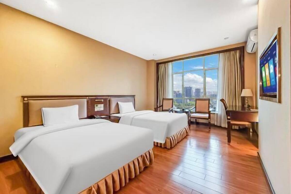 A hotel room with two beds, a desk, a chair, and a large window with a city view.