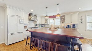 Une cuisine avec des meubles blancs, un îlot central en bois doté de tabourets, des luminaires suspendus et un four encastré.