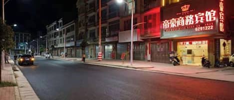 A street view at night with illuminated storefronts, including a hotel with a prominent sign.