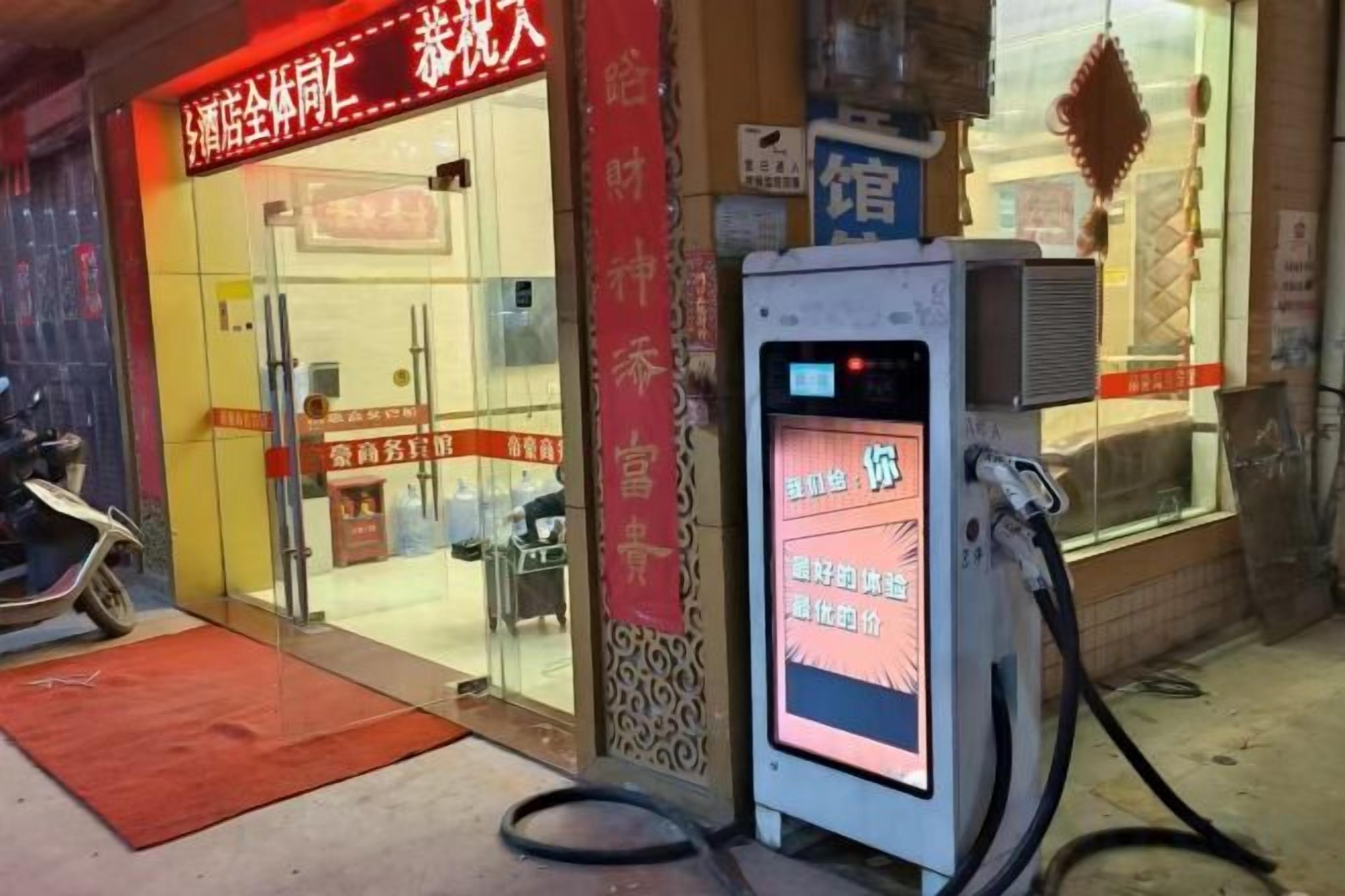 A fuel pump with a digital display and a red mat in front of a building with a glass entrance.