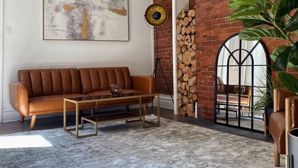 A cozy living room with a brown leather sofa, a coffee table, a modern chandelier, a large abstract painting, and a brick wall with a wooden log display. - Free Parking•Historic Charmer completely renovated (Pittsburgh)