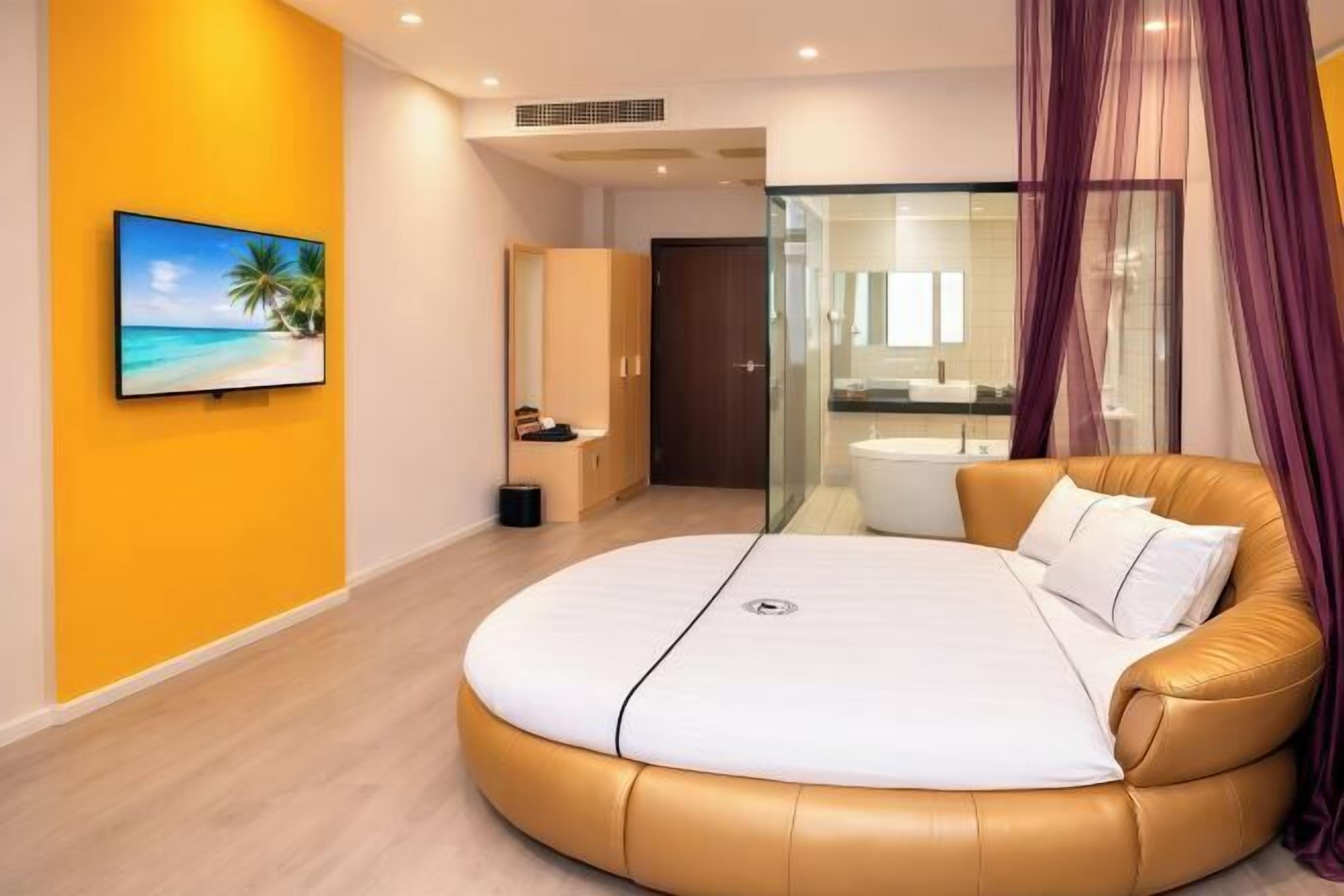 A modern bedroom with a curved bed, a bathroom with a glass shower enclosure, and a tropical-themed painting on the wall.
