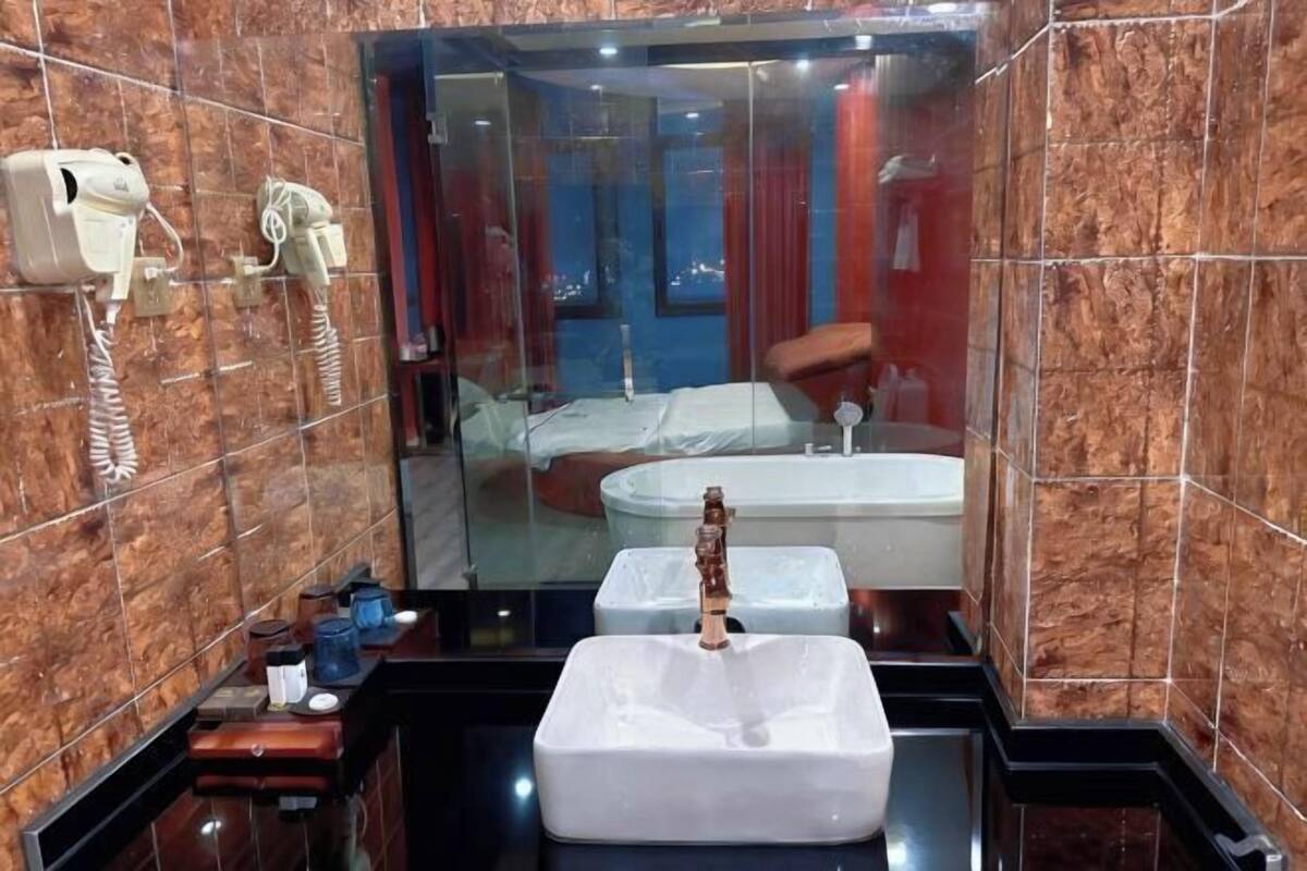 A modern bathroom with a large mirror, a white sink, a hairdryer, and a bathtub visible through the glass partition.