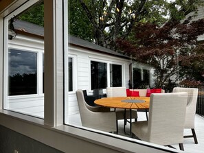 Un espacio moderno para comer al aire libre con una mesa redonda y sillas, ventanas amplias y vistas a un árbol y una casa.