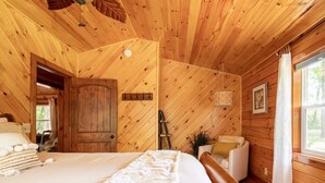 A wooden cabin bedroom with a bed, a chair, a ladder, and a window.
