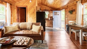 A cozy cabin interior with a wooden ceiling, a sofa with cushions, a dining table, and a kitchen area.