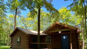 A wooden cabin with a covered porch, a concrete driveway, and a surrounding wooded area.