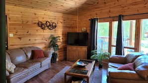 Un interior de cabaña acogedor, con techo de madera, un sofá, una mesa de centro, un televisor y una ventana con cortinas.