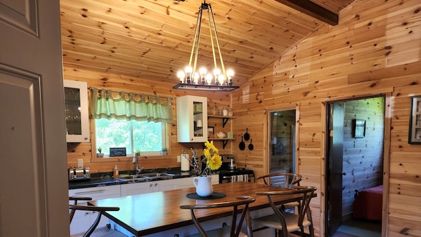 Interior de una cabaña de madera con área de cocina que incluye una isla central, taburetes de bar, una araña y una ventana con cortina verde.