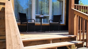 Una terraza de madera con mesas de picnic y dos sillas frente a un amplio ventanal con vistas a un bosque.