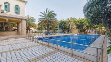A swimming pool with a tiled deck, palm trees, and a building with arched windows.