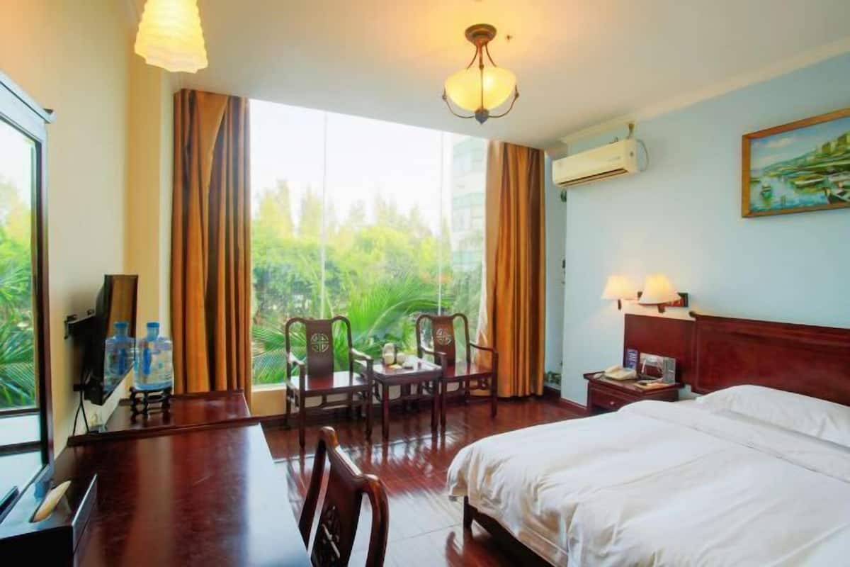 A hotel room with a bed, a desk with a chair, a TV, a window with curtains, and an air conditioning unit.