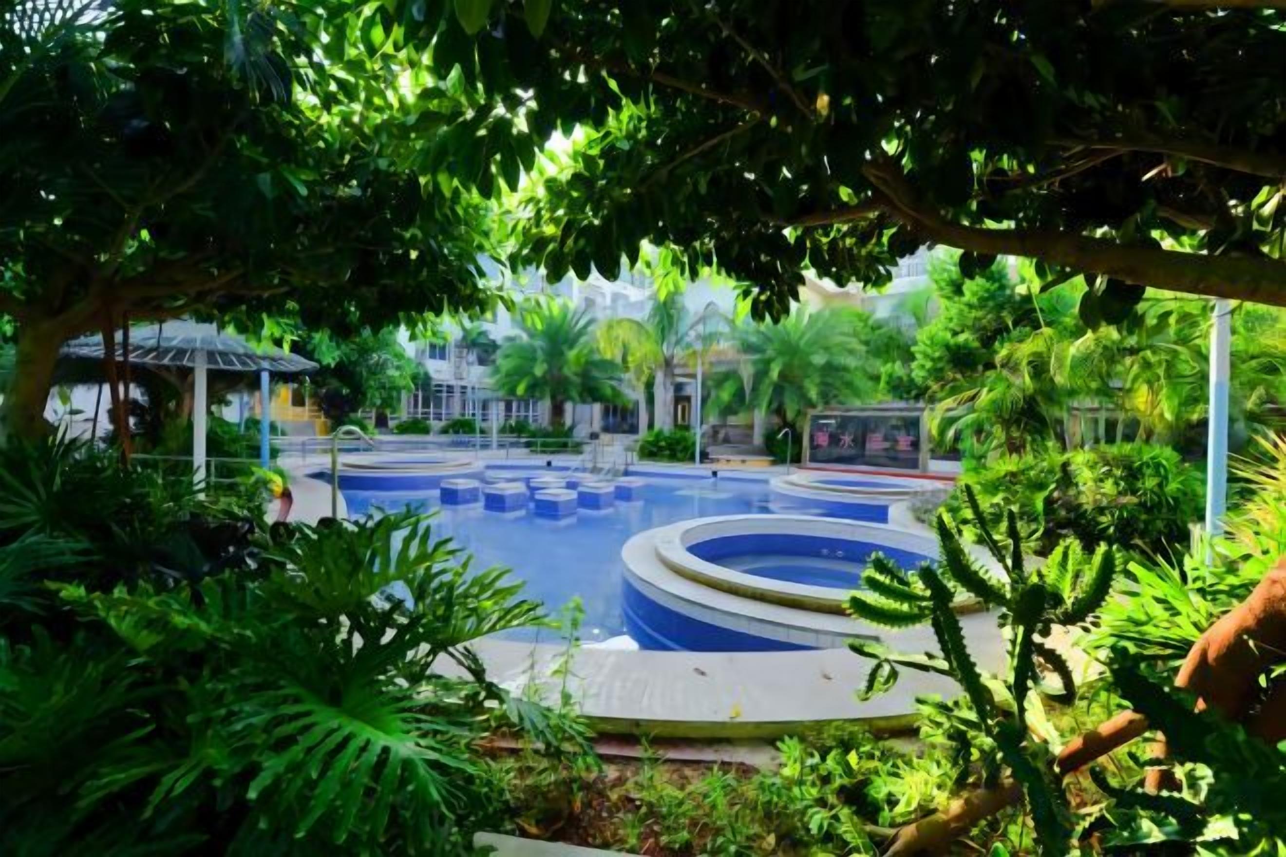 A pool area surrounded by lush greenery, with a gazebo and a small building in the background.