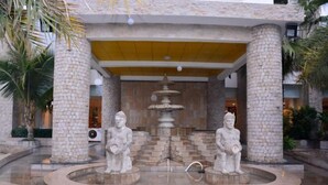 Una fontana in pietra con due statue in un cortile, circondato da palme e un edificio con tetto giallo.