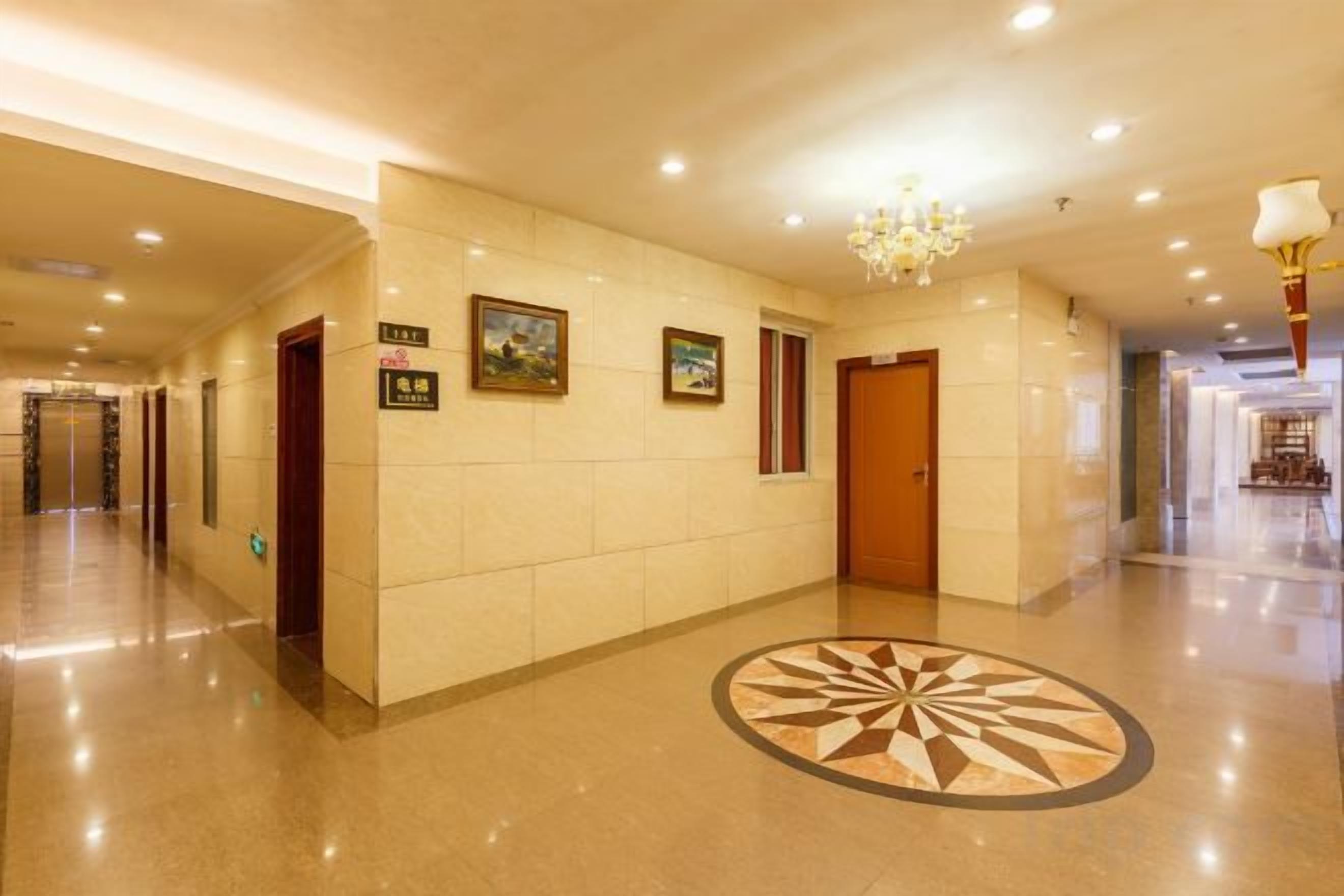 A hotel corridor with a chandelier, framed pictures on the wall, and a patterned floor design.
