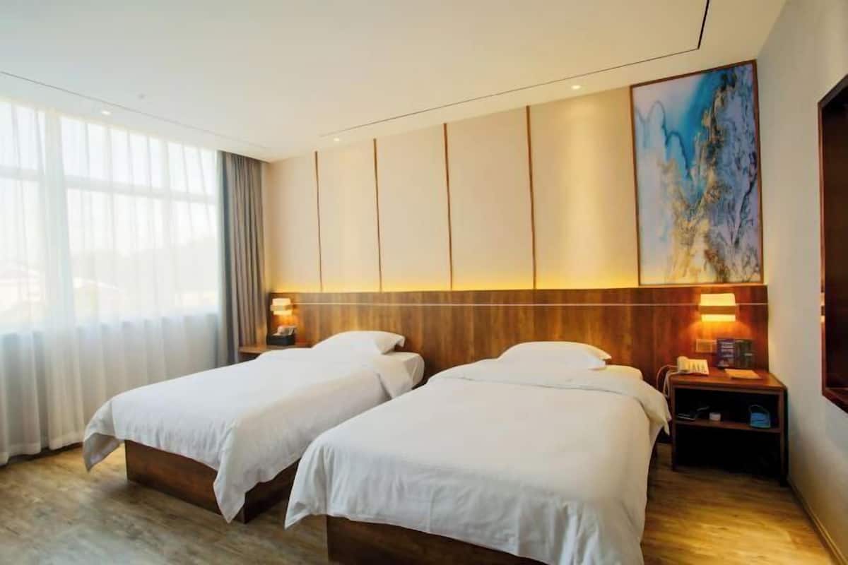 A hotel room with two beds, a large window with curtains, a painting on the wall, and a nightstand with a lamp.