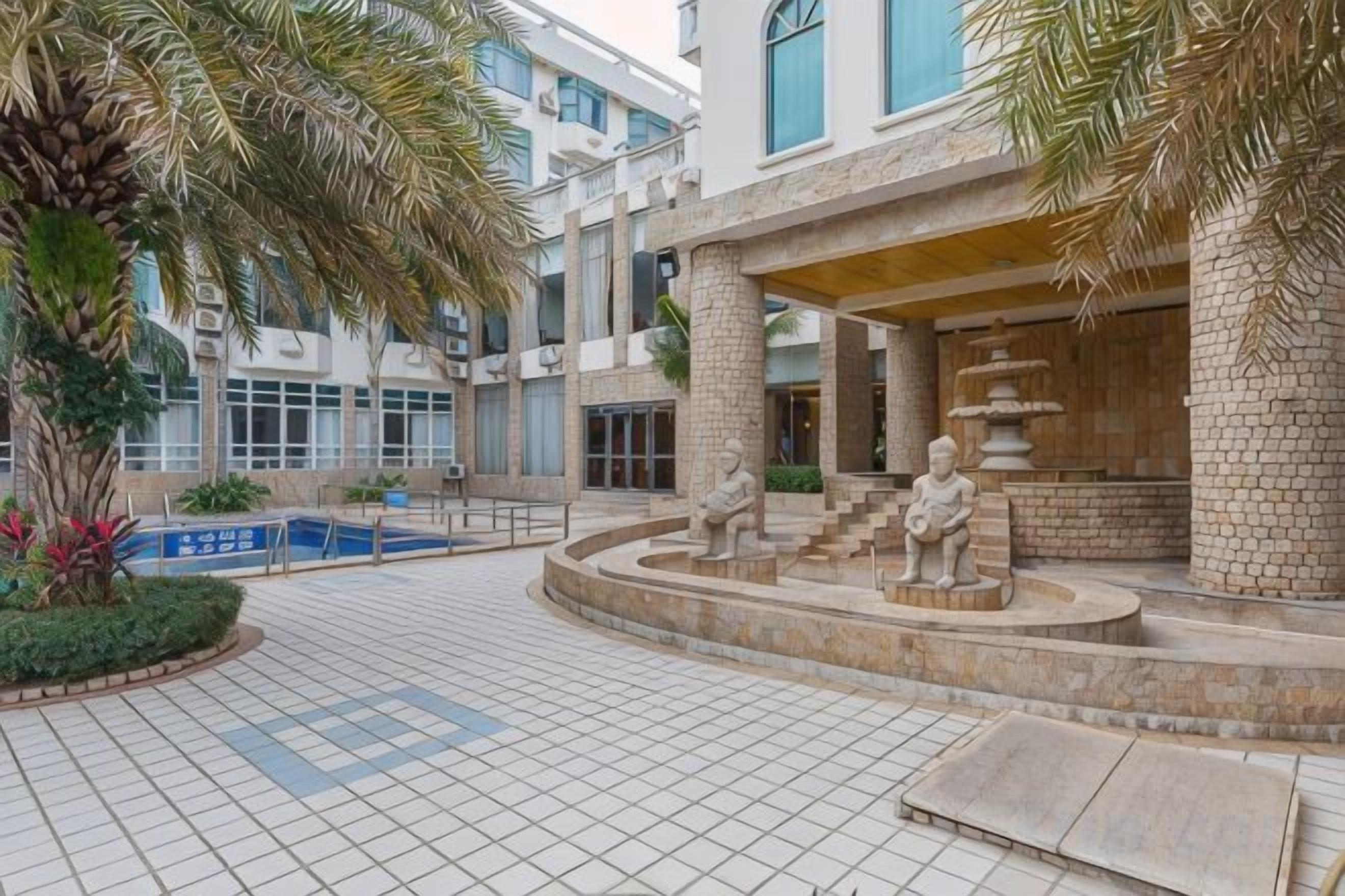 A courtyard with a fountain, palm trees, and a building with a covered area.