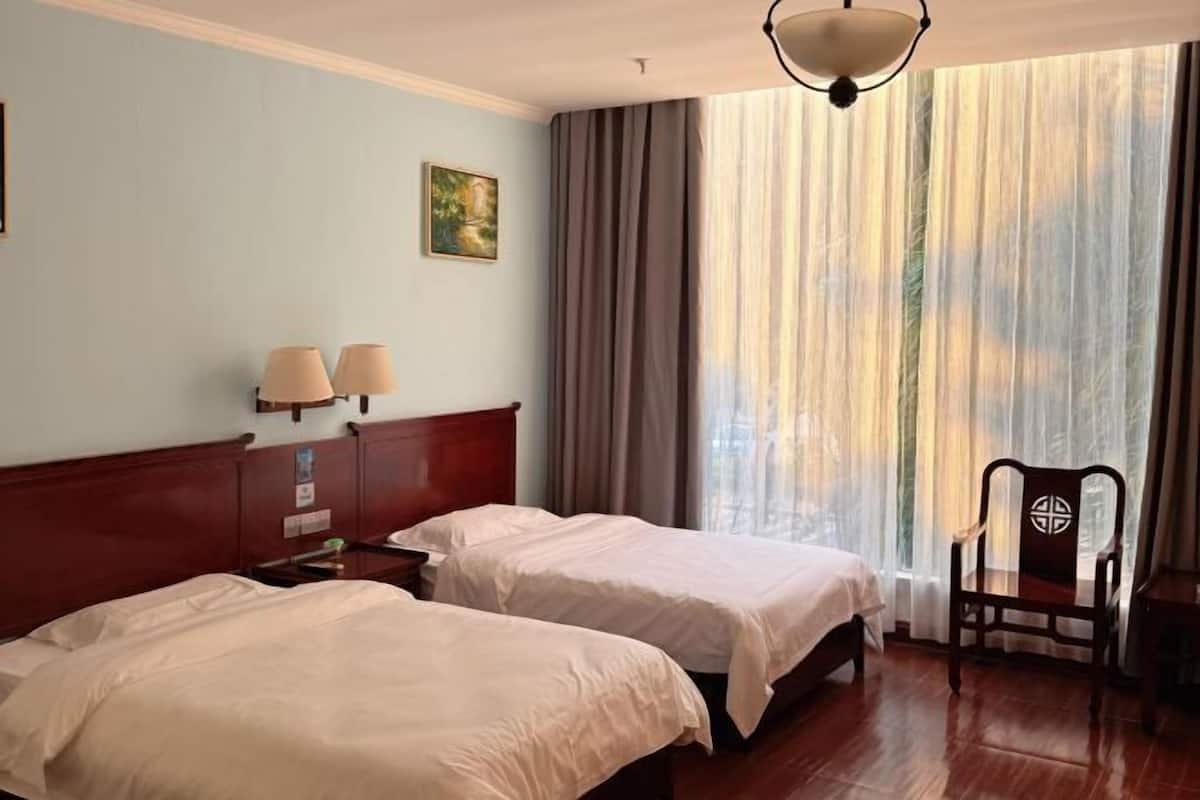 A hotel room with two beds, a wooden headboard, a nightstand, a lamp, a chair, and a window with curtains.