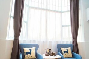 Une pièce avec deux fauteuils bleus, chacun agrémenté d’un coussin orné de la silhouette d’un cerf, une petite table ronde avec une théière et des tasses, et une grande fenêtre avec des rideaux.
