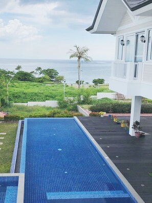 Una piscina con vistas al océano, rodeada de vegetación y un edificio blanco con ventanas y faroles.