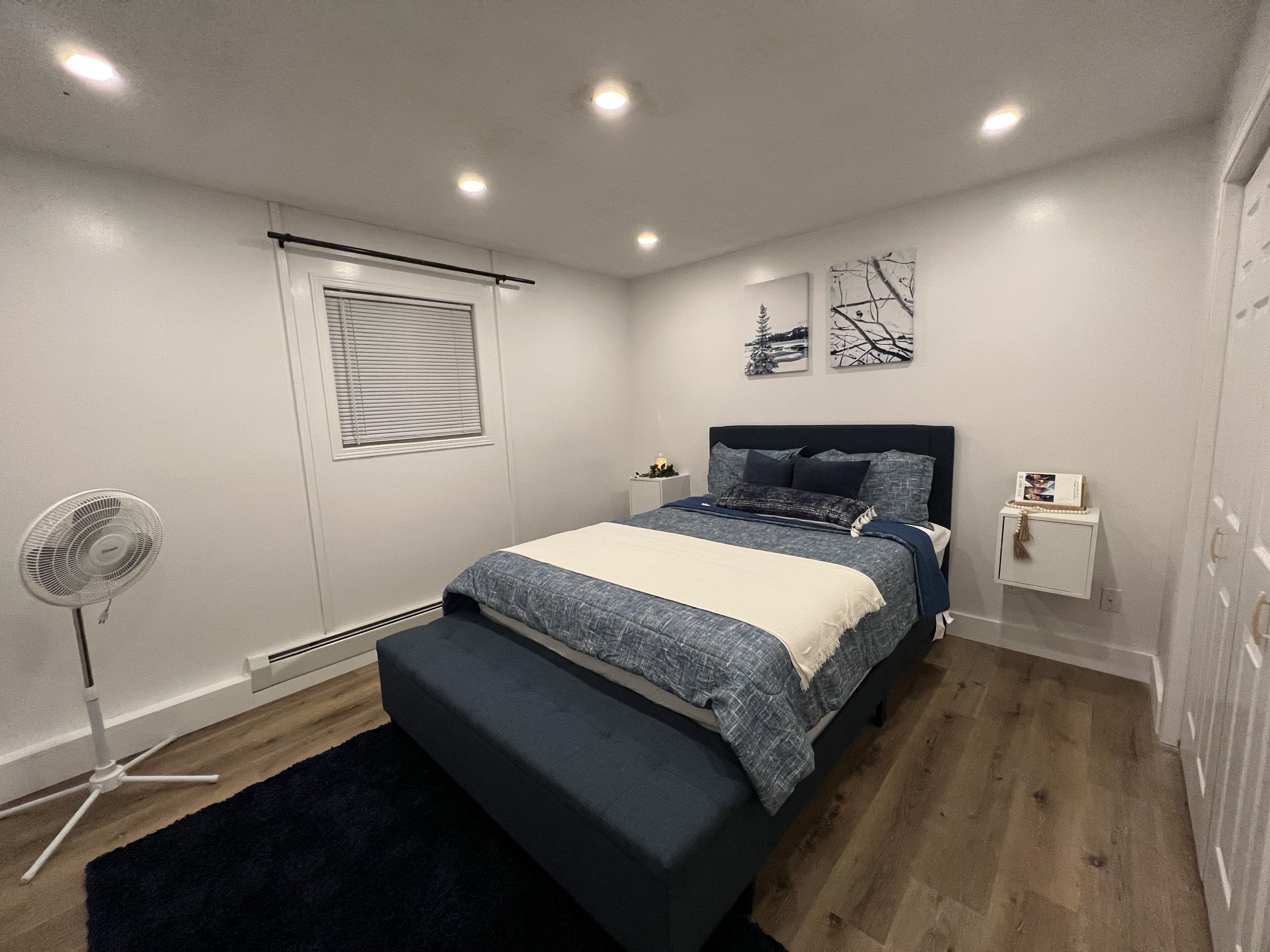 Un dormitorio con cama, mesita de noche, decoración en la pared y un ventilador de pie.
