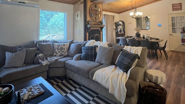 Ein gemütliches Wohnzimmer mit Holzbalkendecke, einem grauen Ecksofa, einem schwarz-weiß gestreiften Teppich und einem Essbereich mit Tisch und Stühlen.